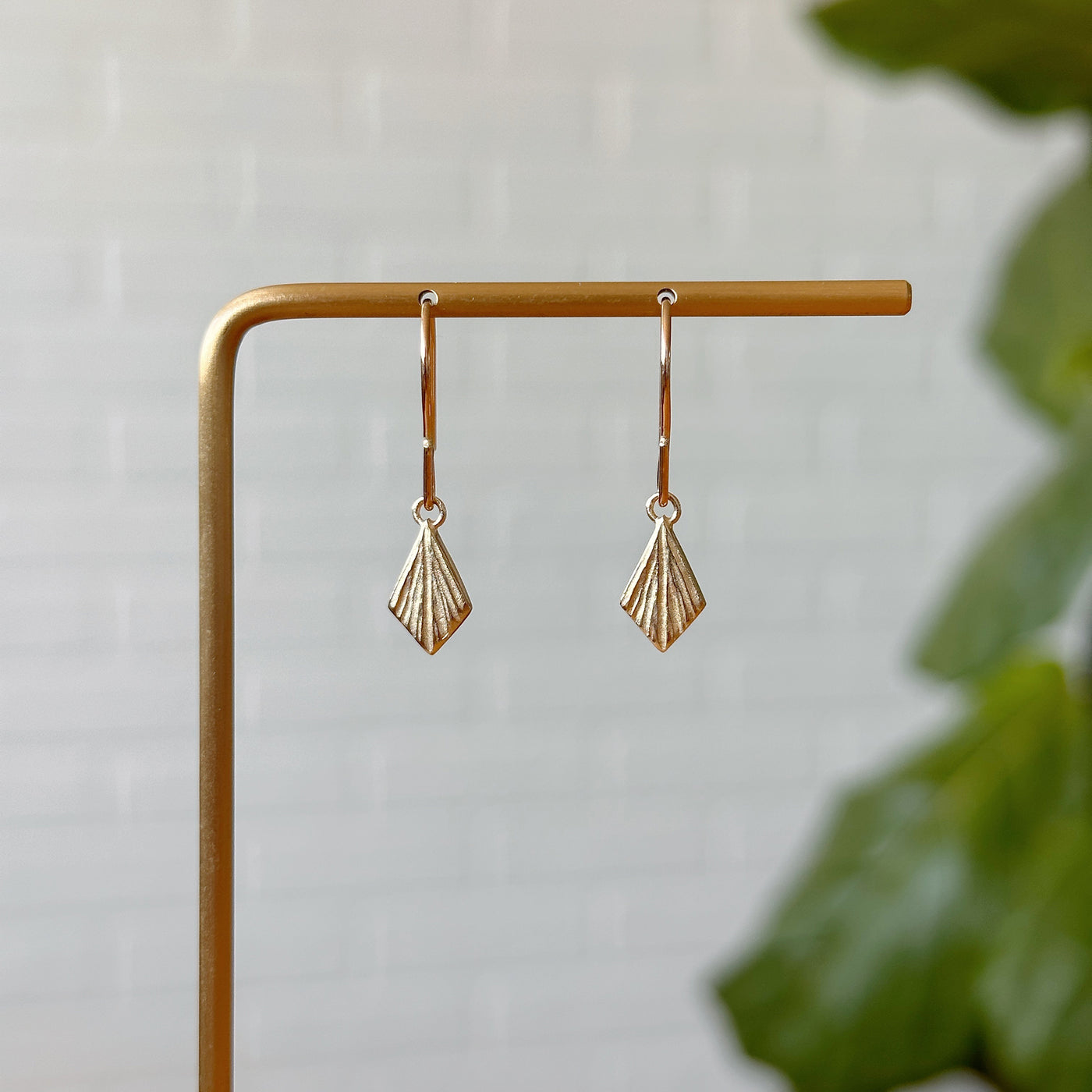 Flame Gold Dangle Earrings hanging in front of a white brick wall, front angle