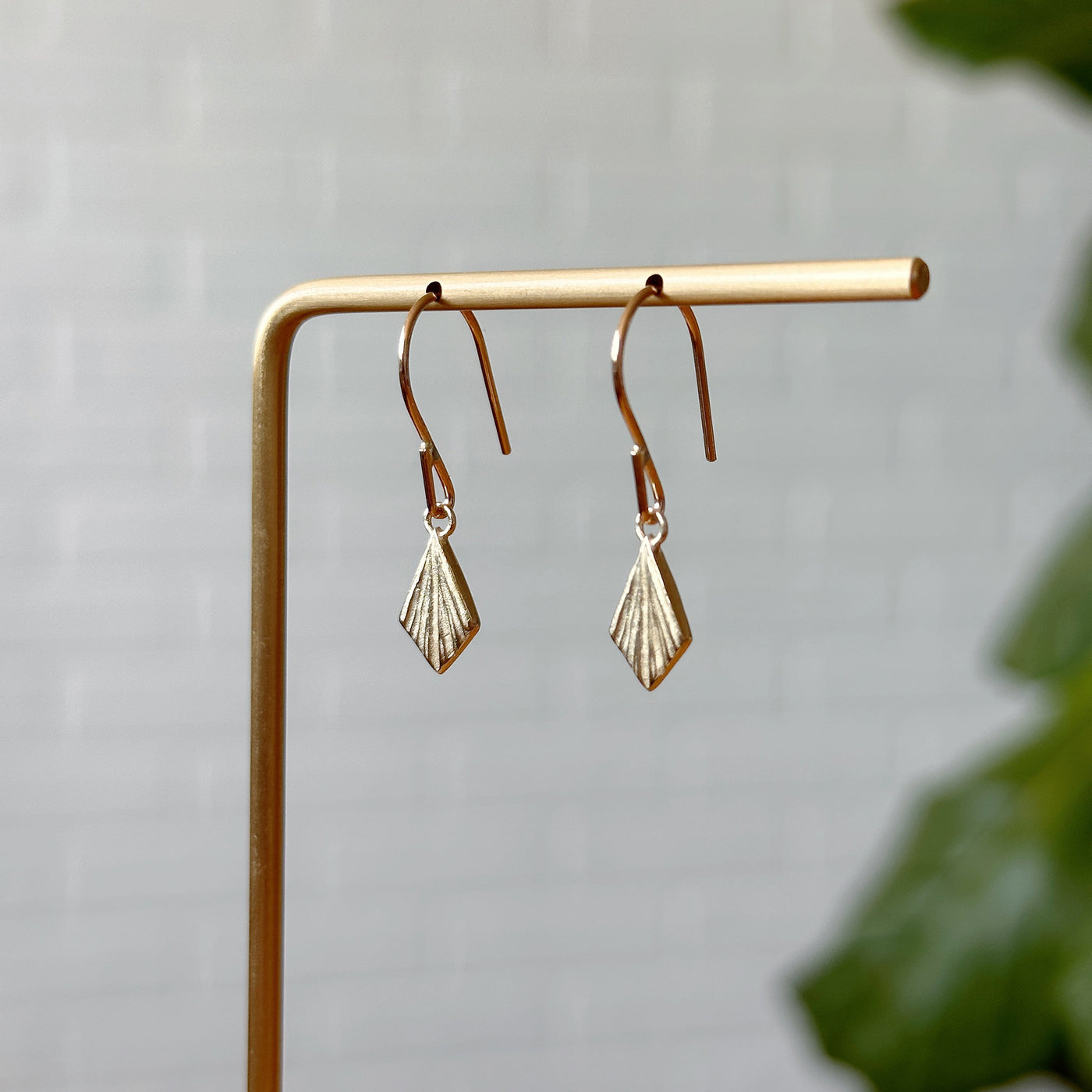 Flame Gold Dangle Earrings hanging in front of a white brick wall, side angle