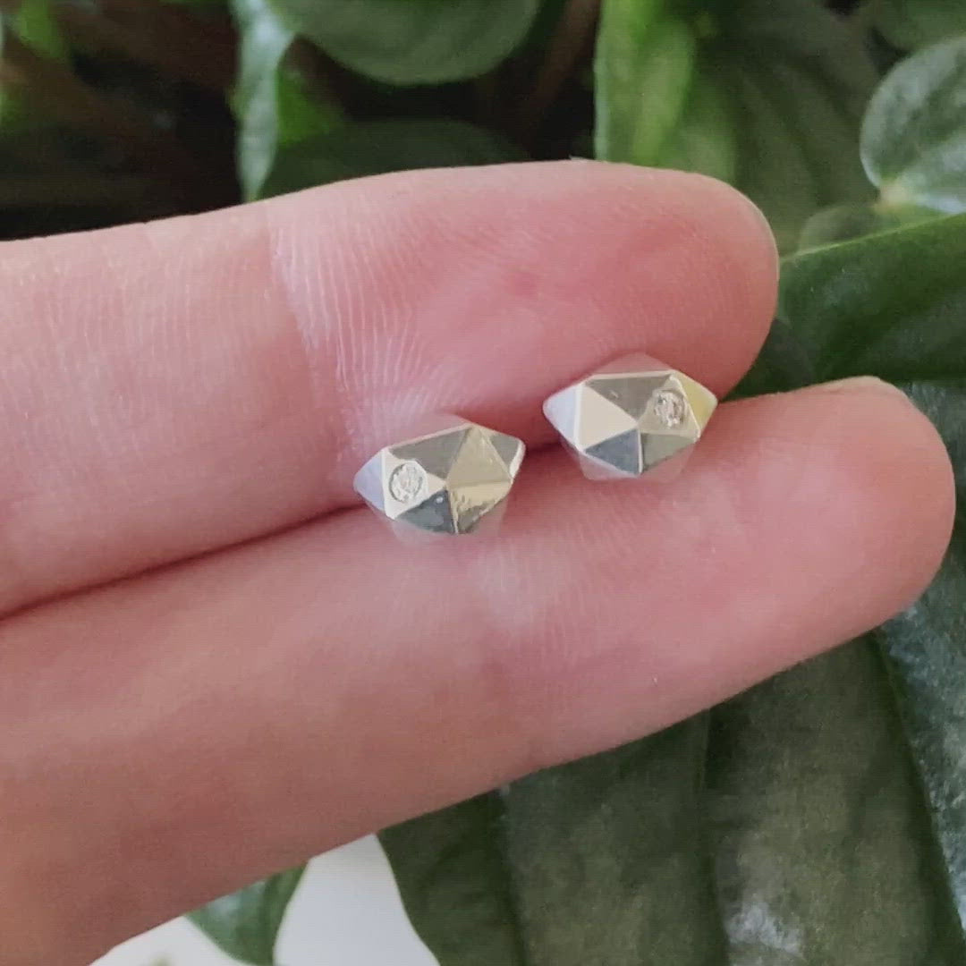 Sterling silver wabi-sabi faceted geometric stud earrings with two flush set diamonds by Corey Egan between two fingers