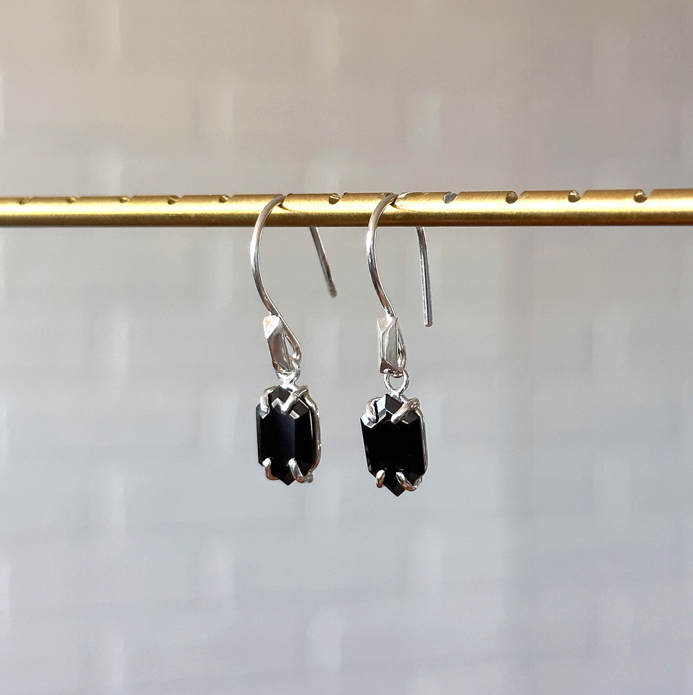 Eloise Black Garnet Earrings in Silver hanging in front of a white brick wall, side angle