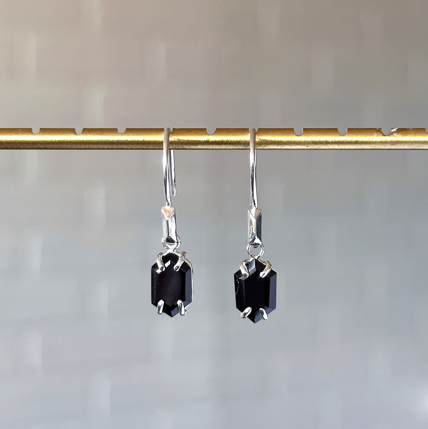 Eloise Black Garnet Earrings in Silver hanging in front of a white brick wall, front angle