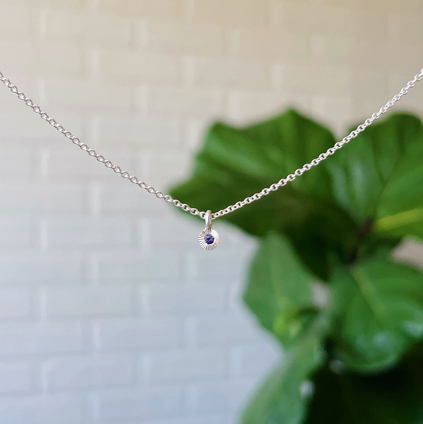 Purple Sapphire Rise Necklace in Sterling Silver hanging in front of a white brick wall