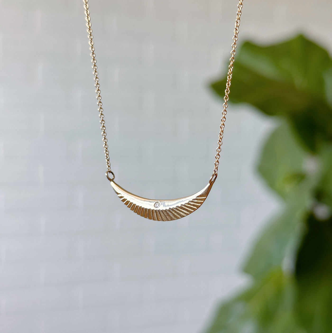 Gold and Diamond Icarus Necklace hanging in front of a white wall, side angle