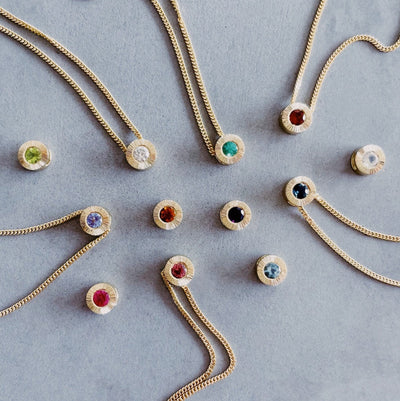 scattered 14k yellow gold aurora pendants with birthstones. Garnet for January,  amethyst for February, aquamarine for March, diamond for April, emerald for May, moonstone for June, ruby for July, peridot for August, blue sapphire for September, pink tourmaline for October, citrine for November, and tanzanite for December
