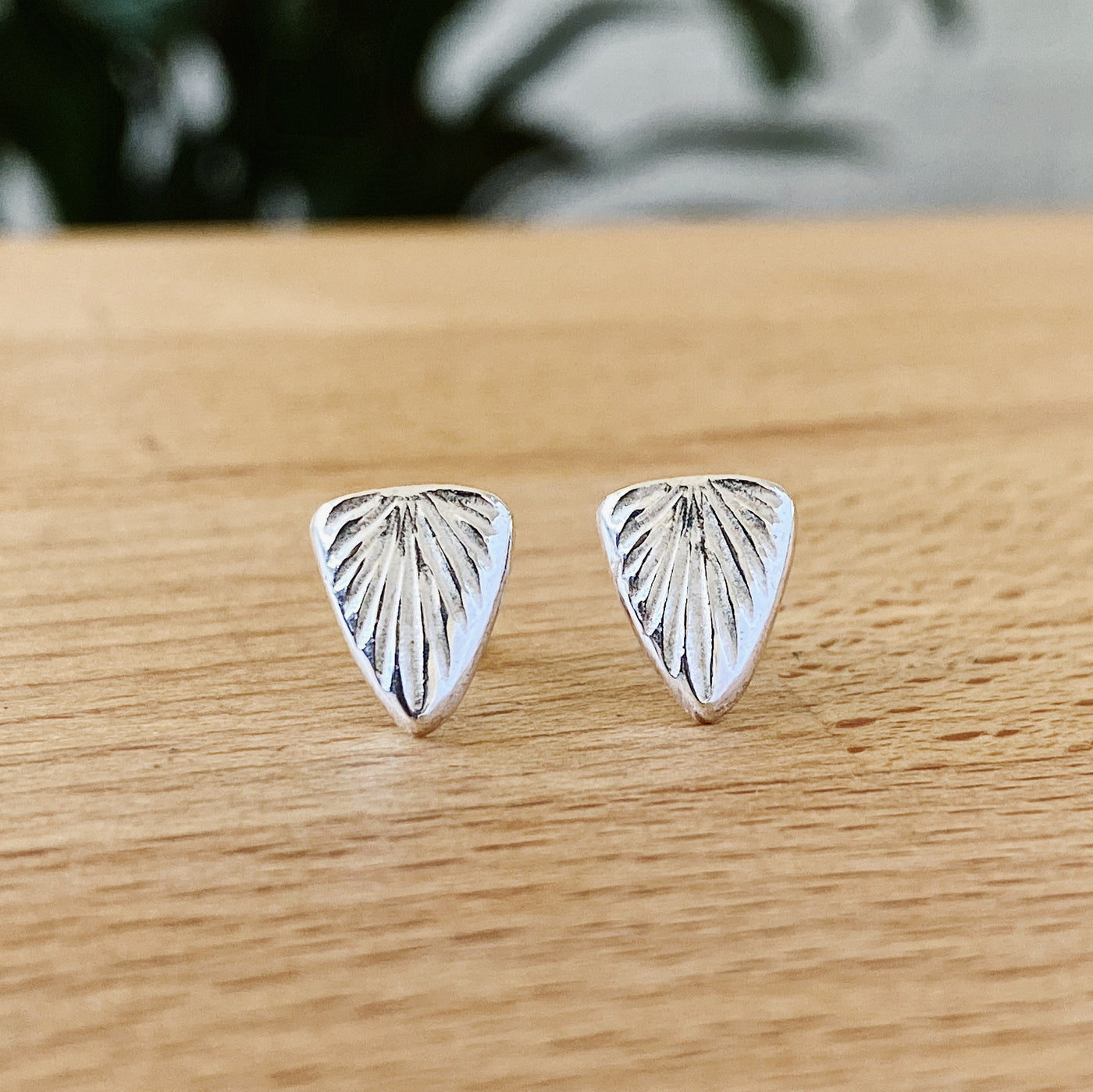 Sterling silver triangular stud earrings with a carved sunburst texture by Corey Egan on a tabletop