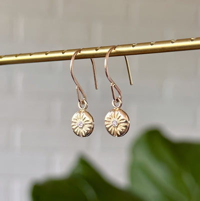Small Lucia Gold Dangle Earrings hanging in front of a white wall, side angle