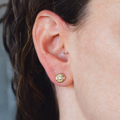 Small round carved sunburst stud earrings with diamond centers in gold on an ear