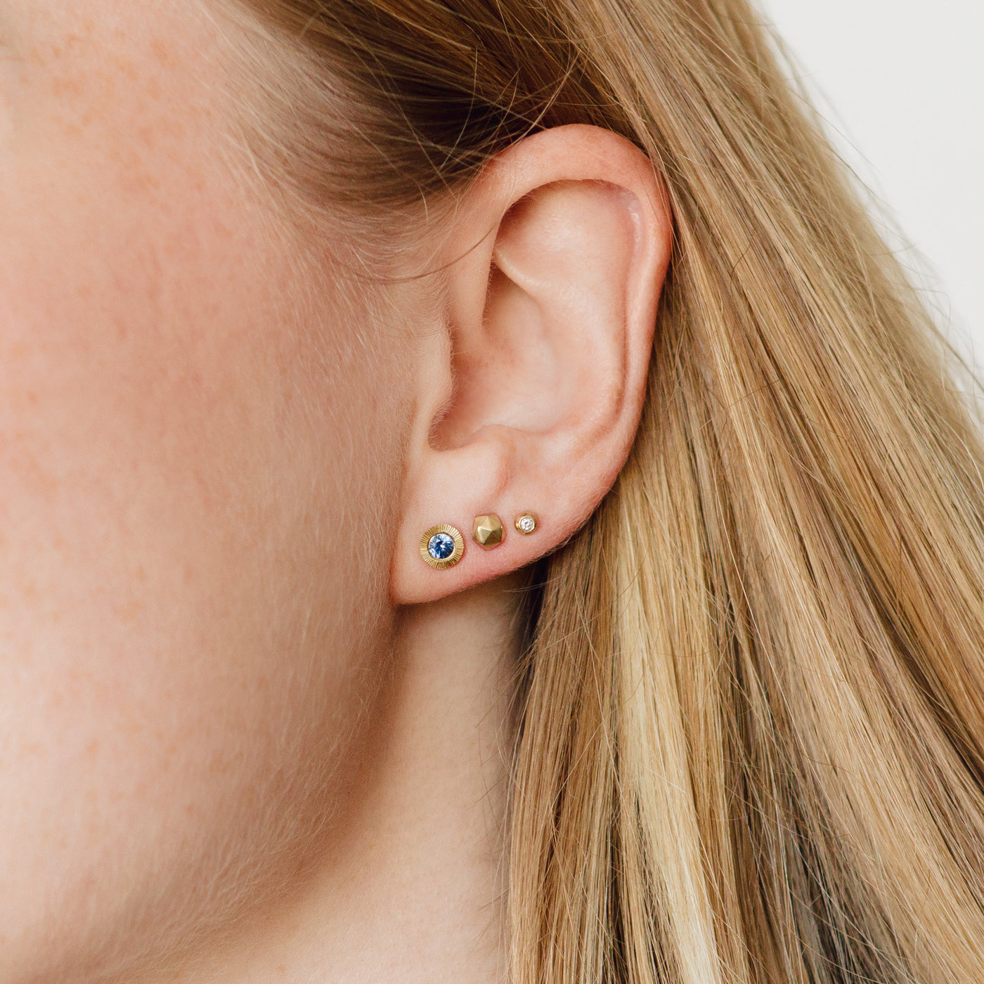 A blue sapphire Aurora Stud, gold micro fragment stud, and diamond and gold droplet stud on an ear