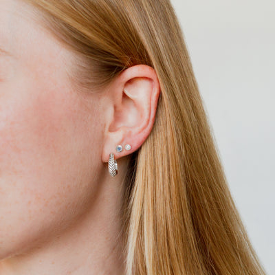 Silver Herringbone hoop earrings paired with a small aurora stud earring with a blue yogo sapphire center and a micro aurora stud earring with diamond center on an ear. 