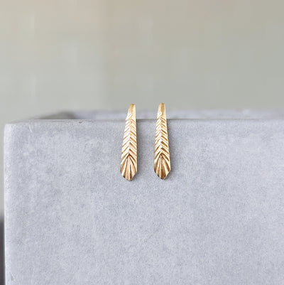 Yellow Gold Tapered Herringbone Stud Earrings hanging on a concrete wall, front facing