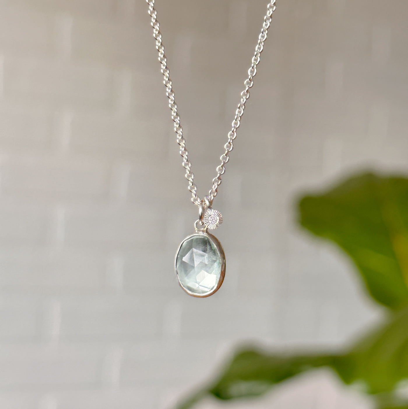 Aquamarine and diamond necklace with silver bezel hanging from a chain in front of a white wall, front angle