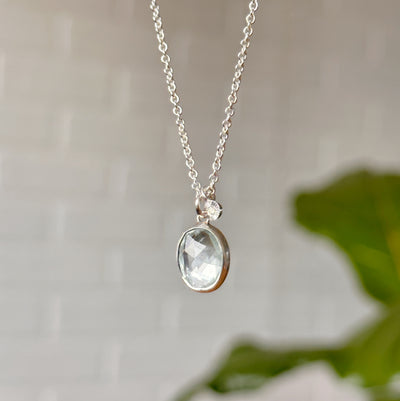 Oval aquamarine pendant necklace in silver in front of a white wall, side angle