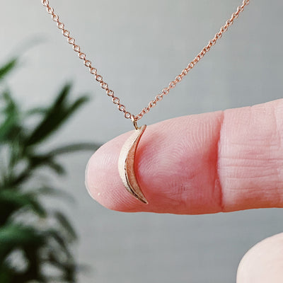 Rose Gold Small Wisp Moon Necklace by Corey Egan on a fingertip for scale