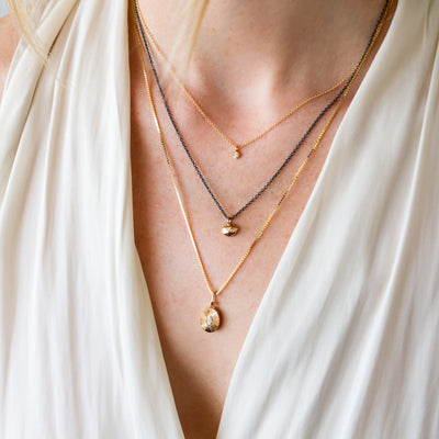 Gold tiny fragment pendant on an oxidized silver chain layered with two other gold necklaces around a neck