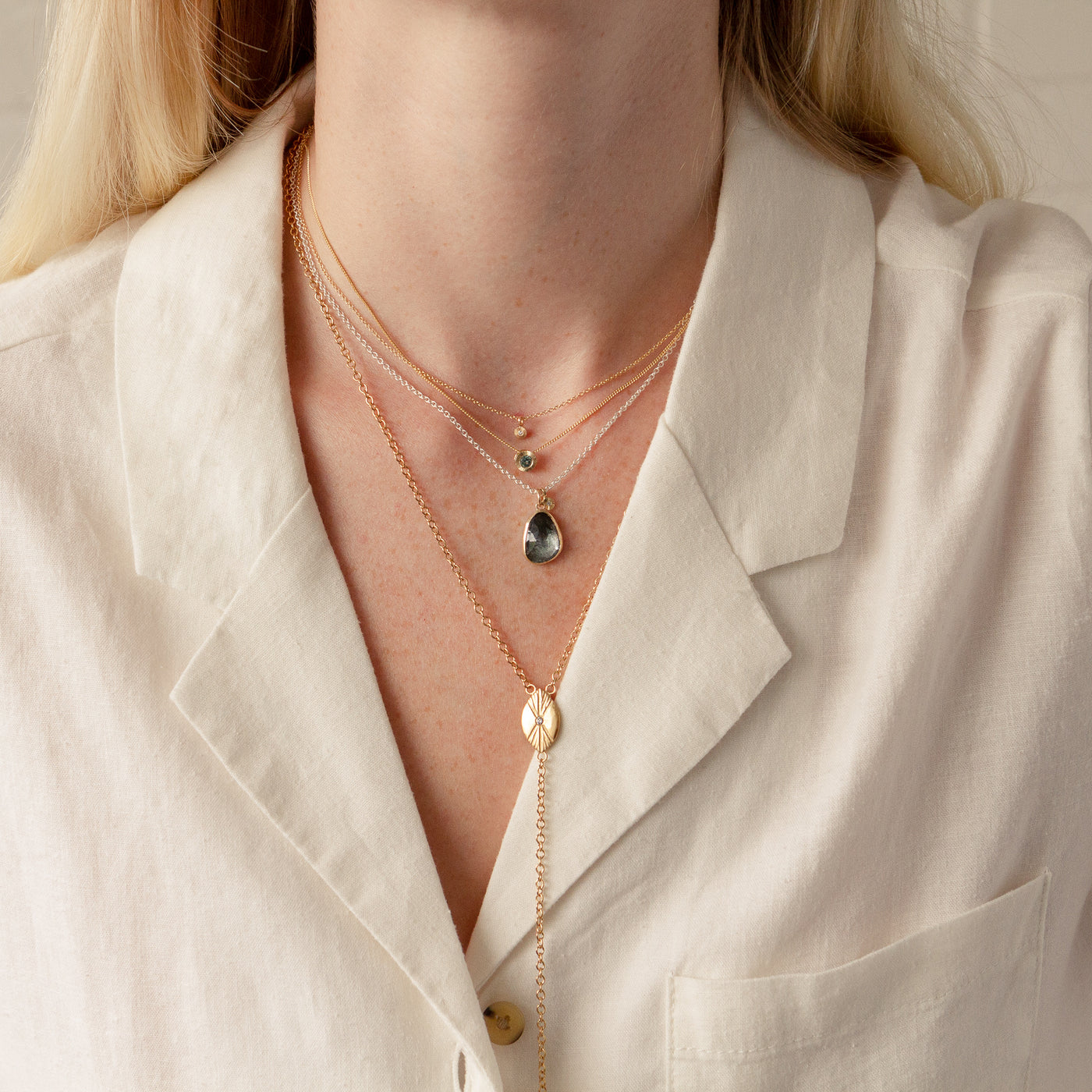 A model wearing a vermeil prism lariat paired with a moss aquamarine theia necklace, a small aurora necklace with teal montana sapphire, and a gold rise necklace on top. 