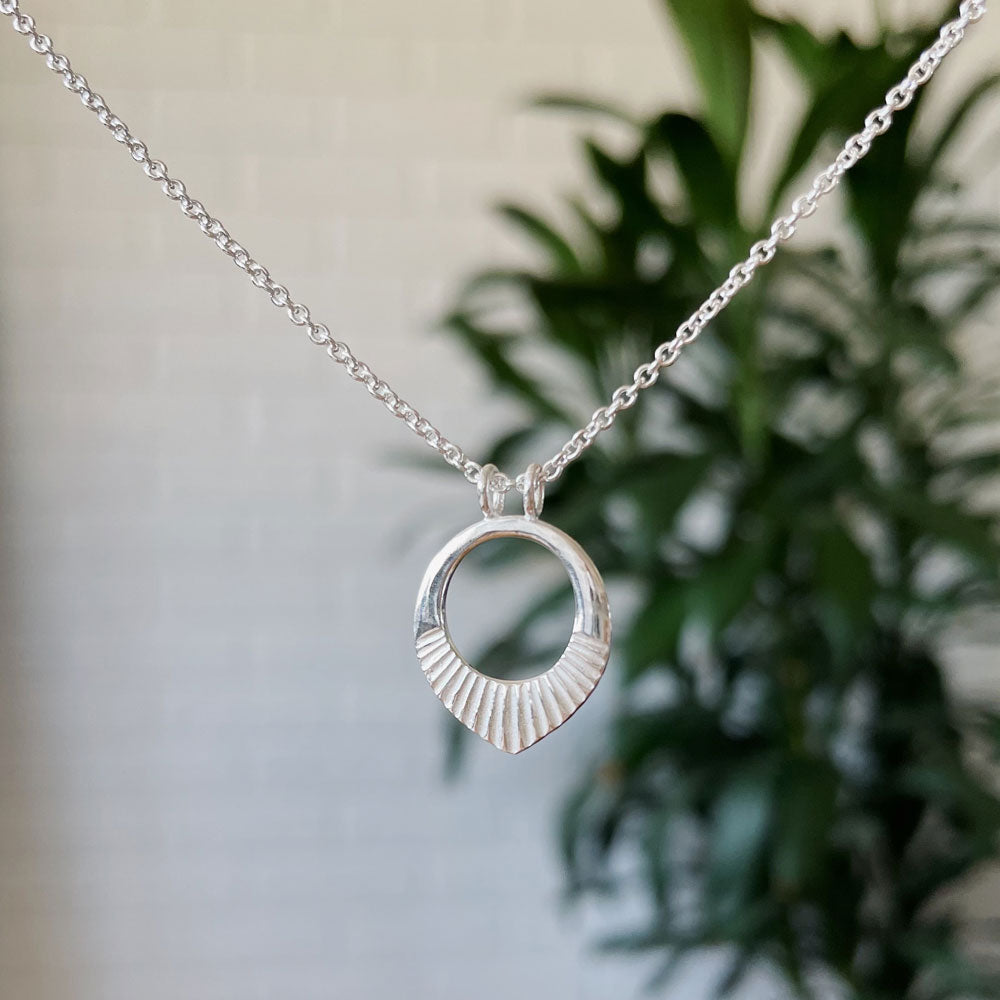 Small silver open petal shape necklace with carved rays across the bottom on natural light