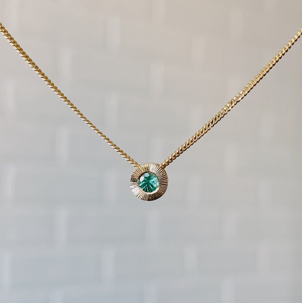 May birthstone 14k yellow gold Aurora necklace with emerald center and engraved sunburst halo border.