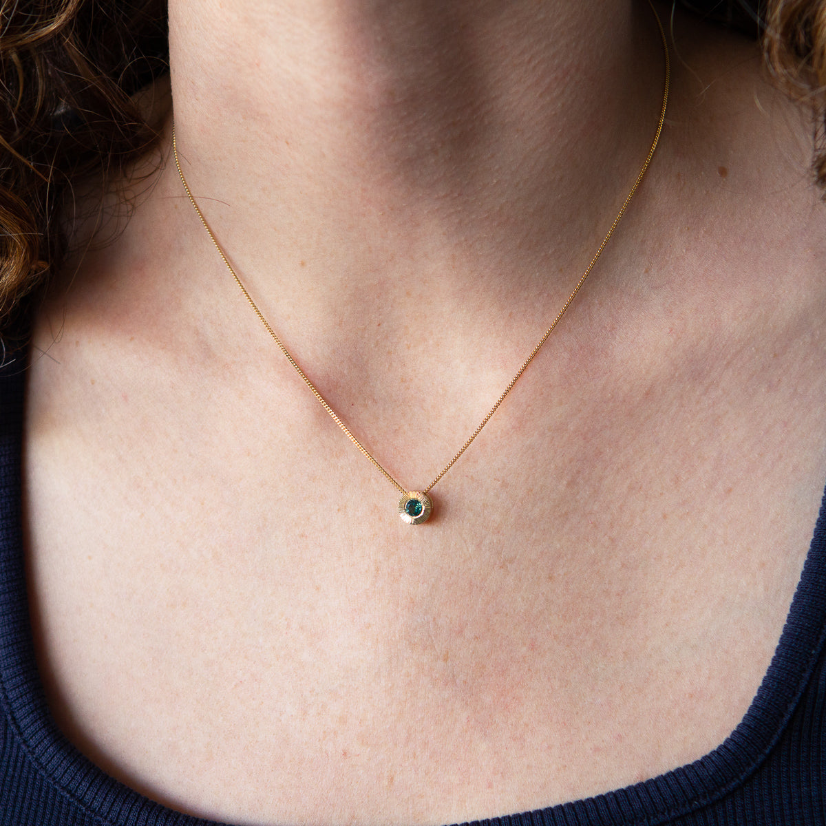 14k yellow gold medium Aurora necklace with a round teal green indicolite tourmaline center and engraved rays halo border on a 16" chain around a neck