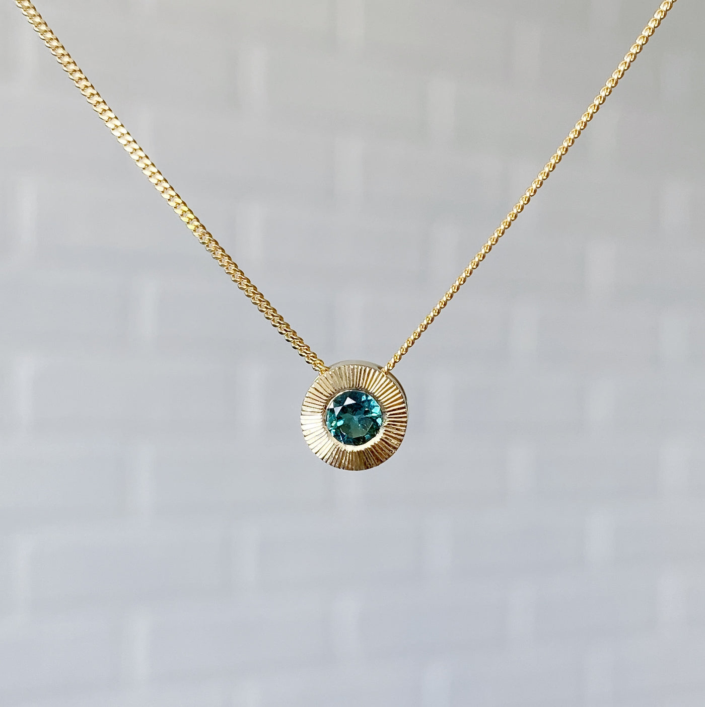 14k yellow gold medium Aurora necklace with a round teal green indicolite tourmaline center and engraved rays halo border in natural light
