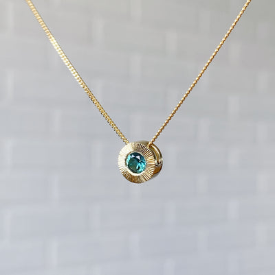 Side view 14k yellow gold medium Aurora necklace with a round teal green indicolite tourmaline center and engraved rays halo border in natural light