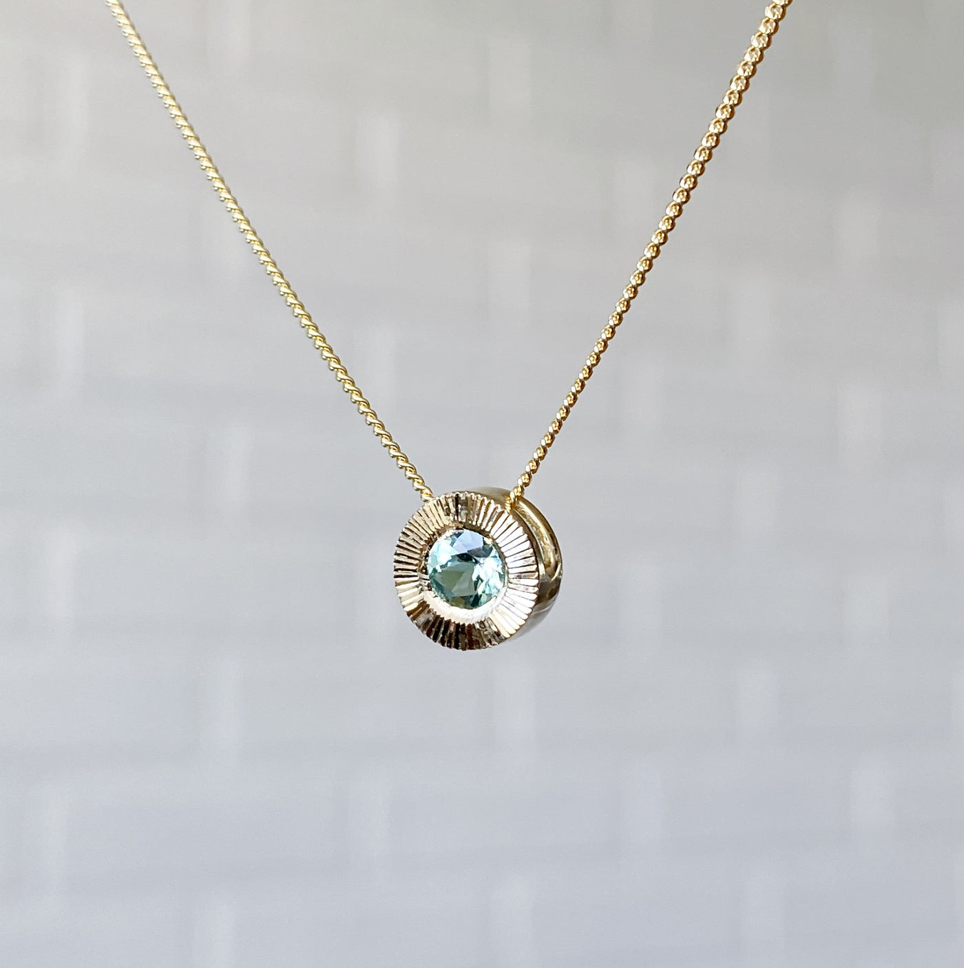 Side view 14k yellow gold medium Aurora necklace with a round seafoam green tourmaline center and engraved rays halo border in natural light