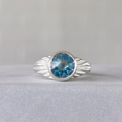 Blue Topaz Silver Sunburst Ring sitting in front of a white wall, front angle