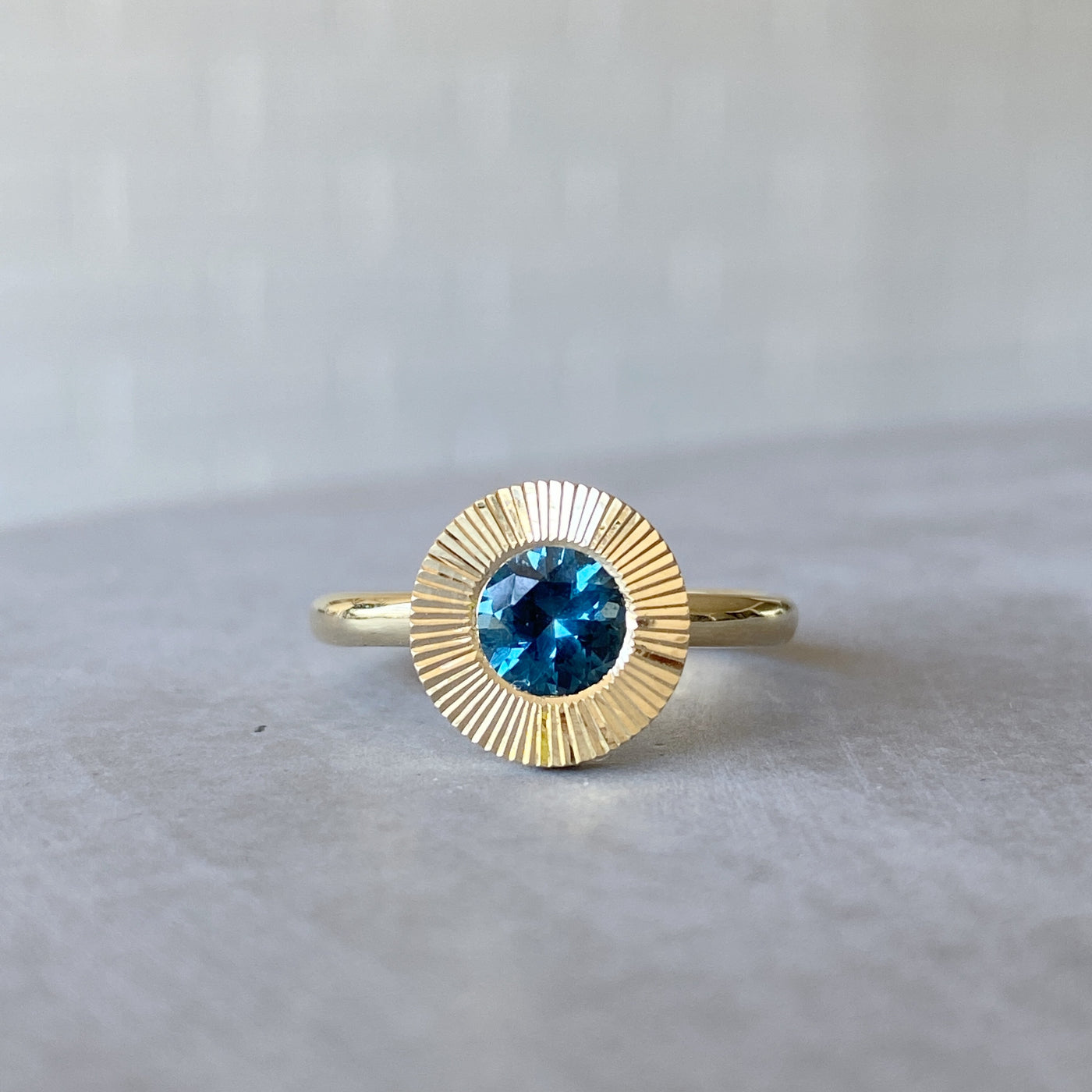 Round Teal Montana sapphire in a 14k yellow gold Aurora ring with an engraved halo border in natural light