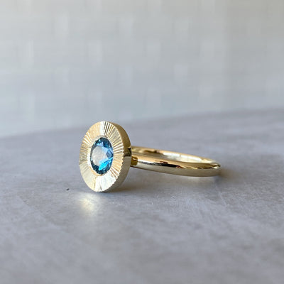 Side view of round teal Montana sapphire in a 14k yellow gold Aurora ring with an engraved halo border in natural light